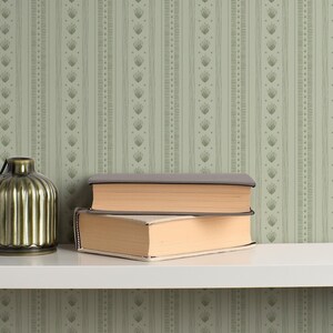 May include: A decorative scene featuring a gold-colored, ribbed vase, and two books with beige pages and gray covers. The items are on a white shelf against a green wallpaper with a vertical pattern.
