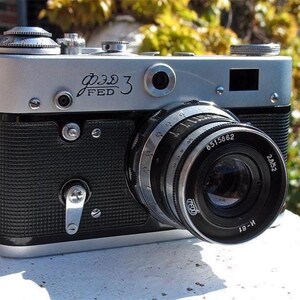 May include: A silver and black FED 3 rangefinder camera featuring a textured black body, a silver top plate with Cyrillic and English text, and a black lens with the markings 8515862 and 2.8/52. The camera includes a manual film advance lever.