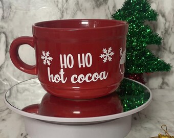 May include: A red ceramic mug with the words "HO HO hot cocoa" in white lettering, and snowflake and cocoa illustrations. A small green Christmas tree with a gold star sits behind the mug. The mug is on a white rotating stand.