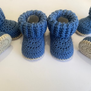 May include: A pair of blue crocheted baby booties with a white trim. The booties have a fold-over cuff and are made with a textured stitch.