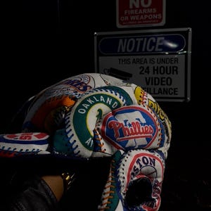 Könnte beinhalten: Baseballkappe mit Aufnähern verschiedener Teams, darunter die Oakland Athletics, Boston Red Sox und Philadelphia Phillies. Die Kappe ist weiß mit bunten Aufnähern und Nähten. Schilder im Hintergrund zeigen "NO FIREARMS OR WEAPONS" und "NOTICE THIS AREA IS UNDER 24 HOUR VIDEO FILLANCE."