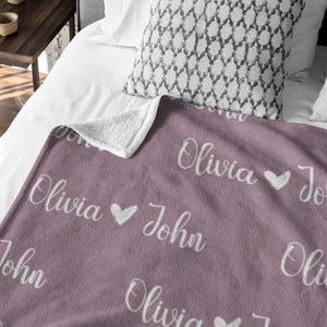 May include: A soft, plush throw blanket with a repeating pattern of the names "Olivia" and "John" in white script with a small heart between each name. The blanket is a dusty rose color.