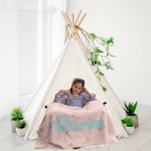 May include: A pink and gold personalized blanket with the name "Olivia" in a teal blue font. A young girl is sitting on a white teepee with the blanket draped over her lap.