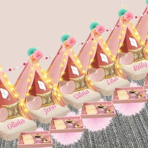 May include: Five pink teepees with gold glitter accents and white lights. Each teepee has a pink heart-shaped pillow and a gold pillow. The teepees are decorated with pink and white pom-poms and a string of pink and white beads. Each teepee has a name on it: Olivia, Jena, Silvia, Lisa, and Reilly. There are pink trays with snacks in front of each teepee.