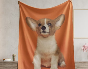 May include: A soft orange fleece blanket featuring a printed image of a brown and white corgi puppy with large ears sitting on a bright orange background.