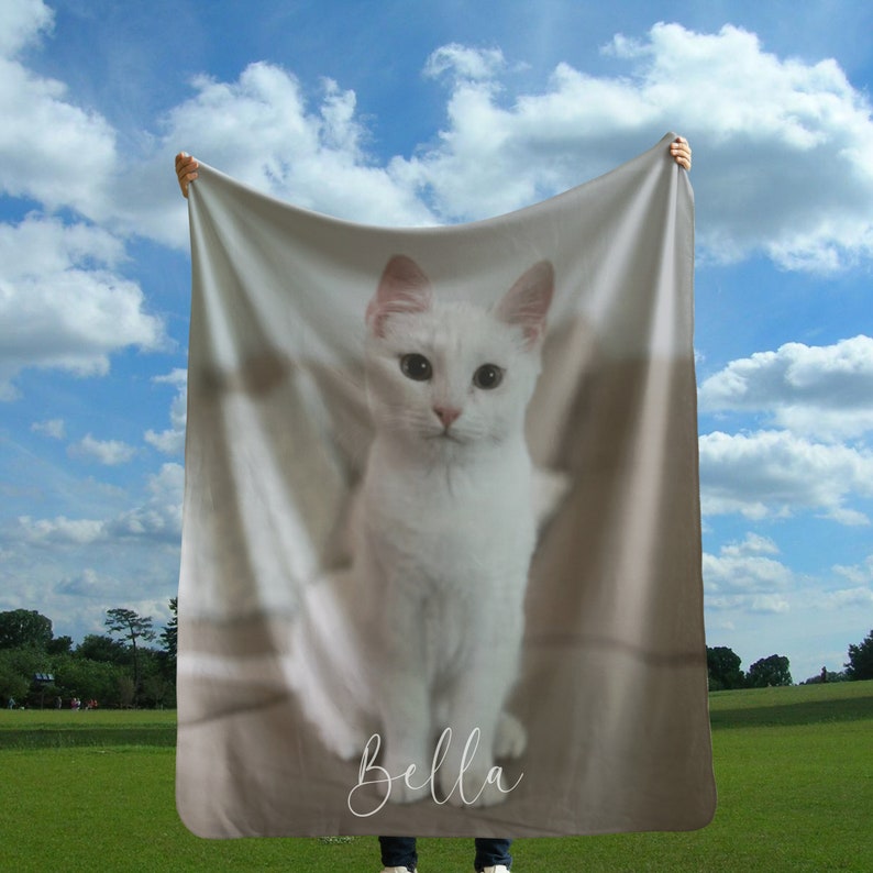 May include: A soft, beige fleece blanket with a white cat printed on it. The cat is looking at the camera and the blanket has the name "Bella" printed on it in white cursive.