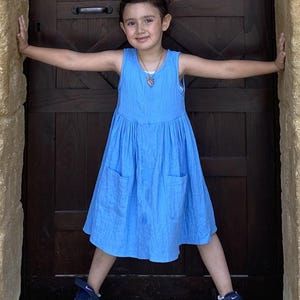 Puede incluir: Un vestido azul sin mangas confeccionado en tejido texturizado, con cintura fruncida y dos bolsillos delanteros de parche. El vestido presenta un cuello redondo sencillo y un largo hasta la rodilla, ofreciendo un dise&ntilde;o limpio y funcional.