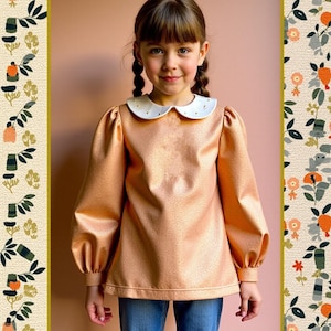 Puede incluir: Una persona joven lleva una camisa de color melocotón con cuello blanco y adornos de perlas. La camisa tiene mangas largas con hombros abullonados y un aspecto ligeramente fruncido. La persona lleva pantalones vaqueros azules.