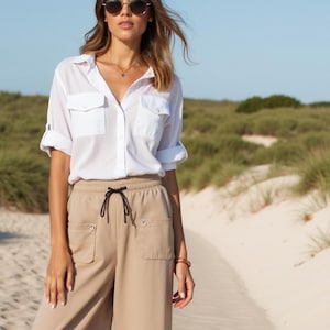 Könnte beinhalten: Eine Frau trägt ein weißes Hemd mit Knöpfen und aufgerollten Ärmeln und eine weite, hellbraune Hose. Sie trägt weiße Turnschuhe und eine Sonnenbrille.