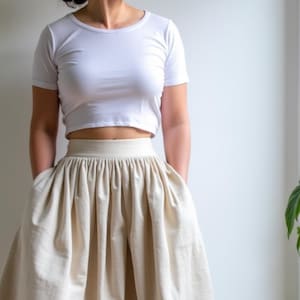 以下が含まれることがあります： 白いクロップドTシャツと、長い、流れるようなクリーム色のスカートを合わせたコーディネート。スカートはハイウエストで、ウエストラインにギャザーが入っています。