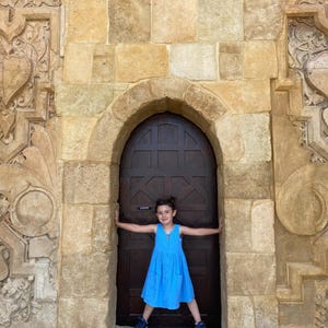 Puede incluir: Un vestido azul brillante sin mangas con cuello redondo y dos bolsillos delanteros, usado por un ni&ntilde;o de pie frente a una puerta arqueada de madera marr&oacute;n oscuro, situada en una pared de piedra color canela con tallas geom&eacute;tricas.