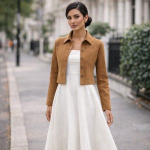 Può includere: Giacca corta in camoscio marrone chiaro con colletto e chiusura con bottoni, indossata sopra un abito bianco. L'outfit è adatto a un'occasione casual o semi-formale.