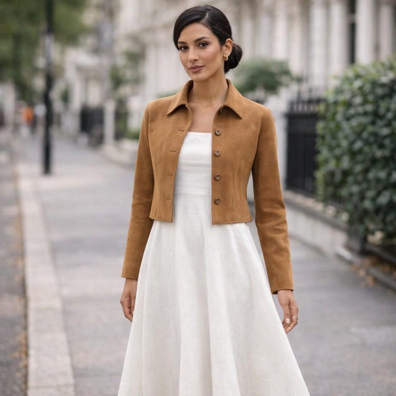May include: A cropped, tan suede jacket with a collared neckline and button closure. The jacket is worn over a white dress. The outfit is suitable for a casual or semi-formal occasion. The jacket's color is a warm, earthy tone.