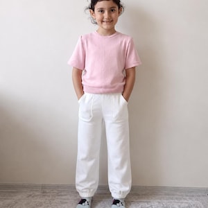 Könnte beinhalten: Eine junge Person trägt einen hellrosa Kurzarmpullover und eine weiße Hose. Die Hose hat Taschen und eine lockere Passform. Die Person trägt außerdem hellblaue Socken mit einem schwarz-weißen Design.