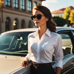 Pode incluir: Uma mulher vestindo uma camisa branca de botões e calças pretas está ao lado de um carro prateado. Ela está usando óculos de sol e tem o cabelo puxado para trás.