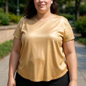 May include: A woman wearing a gold-colored, short-sleeved t-shirt and black pants. The shirt has a smooth, silky texture. The background features a blurred outdoor setting with trees and a paved walkway.