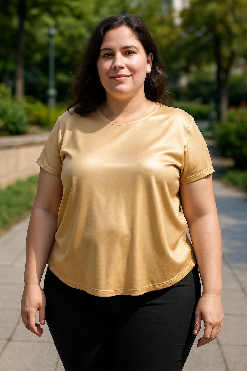 May include: A woman wearing a gold-colored, short-sleeved t-shirt and black pants. The shirt has a smooth, silky texture. The background features a blurred outdoor setting with trees and a paved walkway.