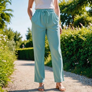 Op de afbeelding: Lichtblauwe broek met een trekkoord in de taille en zakken. De broek heeft een recht model. De persoon draagt een witte top en sandalen. De achtergrond is een pad met groen.