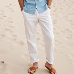 Peut inclure: Une personne portant un pantalon blanc avec les ourlets retroussés et des sandales en cuir marron. La personne est debout sur une plage de sable.