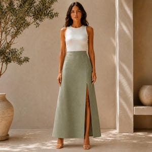 May include: A woman in a white vest top and a sage green maxi skirt with a high slit. The skirt is a solid colour and reaches the floor. The outfit is simple and elegant, suitable for various occasions.
