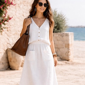 Op de afbeelding: Een vrouw in een witte linnen outfit, bestaande uit een mouwloze top met knopen en een midi-rok met zakken. Ze draagt een bruine leren tas en draagt een zonnebril en sieraden. De achtergrond toont een stenen muur en de zee.