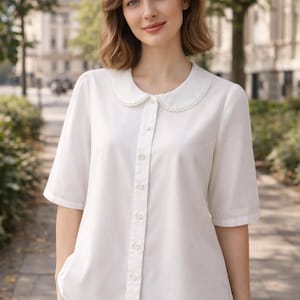 Può includere: Una camicia bianca abbottonata con colletto rotondo e maniche corte. Il colletto ha un bordo di perline delicato. La camicia è abbinata a una gonna beige. Lo sfondo presenta una scena all'aperto sfocata con alberi ed edifici.