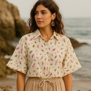 Op de afbeelding: Crèmekleurig overhemd met knopen en een botanische print met roze, groene en bruine bladeren en twijgen. Het shirt met korte mouwen wordt gecombineerd met een bruine broek met trekkoord. Het model draagt gouden oorbellen.