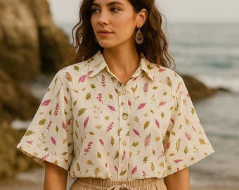 May include: Cream-coloured button-down shirt with a botanical print featuring pink, green, and tan leaves and sprigs. The short-sleeved shirt is paired with tan drawstring trousers. The model is wearing gold earrings.