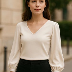 May include: A woman wearing a cream-coloured, long-sleeved v-neck blouse with puffed sleeves and black trousers.