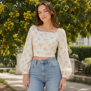 Peut inclure: Un haut court à manches longues de couleur crème avec un col carré et un motif floral. Le haut présente un motif délicat de petites fleurs roses et jaunes. Le modèle porte un jean bleu clair. L'arrière-plan comprend des fleurs jaunes et du feuillage vert.
