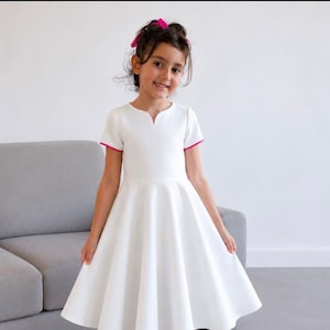 Peut inclure: Une robe blanche à jupe évasée et manches courtes. La robe a une encolure en V et une bordure rose sur les manches. Une jeune personne aux cheveux foncés et un nœud rose sourit à la caméra. La robe semble être faite d'un tissu lisse.