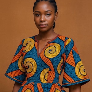 May include: A woman wearing a vibrant, patterned top with a V-neckline and flared sleeves. The top features a blue base with orange and yellow spiral and geometric designs. The neckline is trimmed with red stitching, creating a striking contrast.
