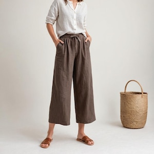 Może przedstawiać: Kobieta w białej koszuli lnianej i brązowych szerokich spodniach lnianych ze sznurkiem w pasie. Stoi na białym tle z brązowym wiklinowym koszem po prawej stronie.