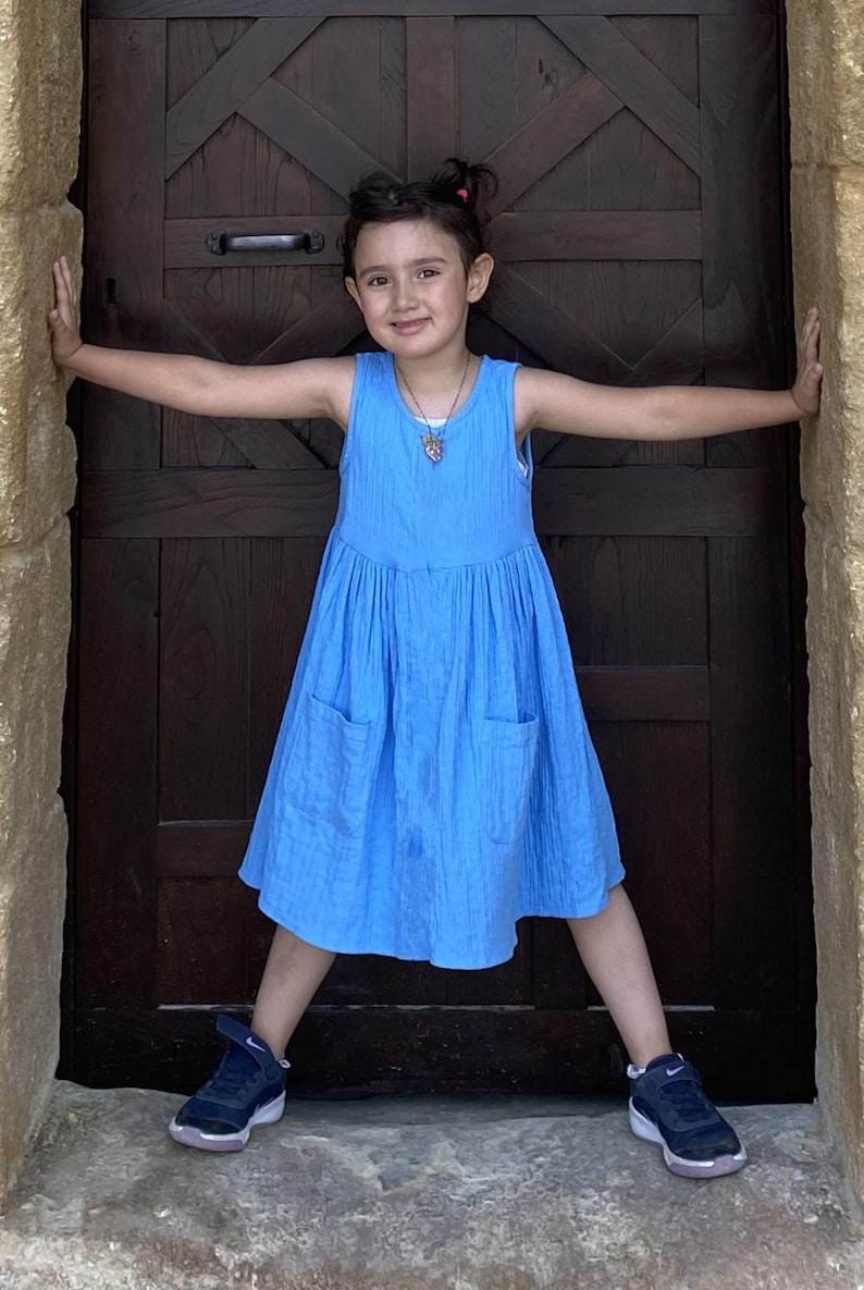 Puede incluir: Un vestido azul sin mangas confeccionado en tejido texturizado, con cintura fruncida y dos bolsillos delanteros de parche. El vestido presenta un cuello redondo sencillo y un largo hasta la rodilla, ofreciendo un dise&ntilde;o limpio y funcional.