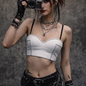 Könnte beinhalten: Weißes Crop-Top mit schwarzem Besatz und Spaghettiträgern. Das Model trägt einen schwarzen Gürtel mit Kettendetails, eine schwarze Jeans und mehrere silberne Halsketten. Eine Vintage-Kamera wird vor das Gesicht gehalten.