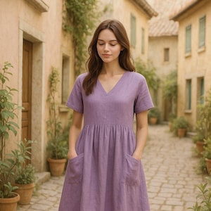 May include: A woman wearing a lavender linen dress with a v-neck and short sleeves. The dress has two pockets and a gathered waist.