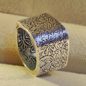 May include: A silver ring with a square face featuring an intricate floral design.
