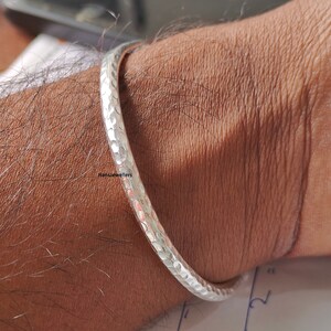 Op de afbeelding: Een zilverkleurige gehamerde metalen armband. De armband heeft een textuur met een reeks kleine, gelijkmatig verdeelde inkepingen. De armband wordt om een pols gedragen.