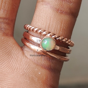 Könnte beinhalten: Ein Set aus drei Ringen aus Roségold mit einem zarten Opal-Edelstein. Die Ringe sind auf einem Finger übereinander gelegt und haben ein geflochtenes Seil-Design.