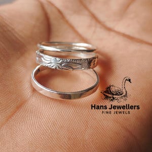 Puede incluir: Tres anillos de plata apilados uno encima del otro. El anillo superior tiene un diseño floral. El anillo inferior es liso. Los anillos están en la mano de una persona. La imagen también incluye el texto "Hans Jewellers Fine Jewels".