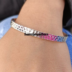 Puede incluir: Un brazalete de plata martillado con un diseño rosa y azul. El brazalete está en la muñeca de una persona.