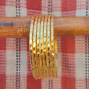 Op de afbeelding: Een set van acht gouden metalen armbanden met een gehamerde textuur. De armbanden zijn op elkaar gestapeld en in een halve cirkel gerangschikt.