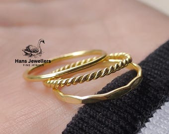 May include: Three gold rings stacked on a finger. The top ring is a simple band, the middle ring is a twisted rope design, and the bottom ring is a hammered band.