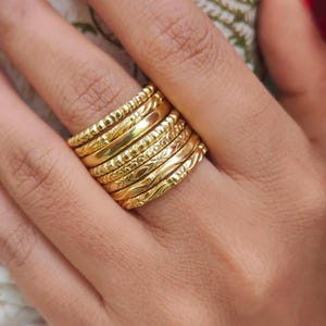 以下が含まれることがあります： ビーズ、エッチング、滑らかなデザインなど、さまざまなテクスチャのゴールドカラーのリングのスタック。リングは指に着用され、さまざまなパターンとジュエリー全体の輝きを披露しています。