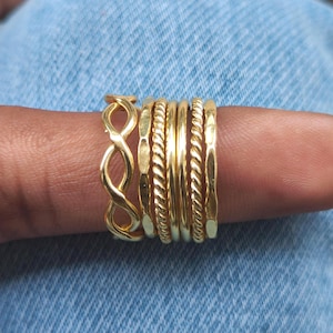 Può includere: Una collezione di anelli dorati con vari design, tra cui intrecci e motivi a infinito, indossati su un dito. Gli anelli sono presentati su uno sfondo di denim blu, evidenziando i dettagli e la lucentezza dei gioielli.