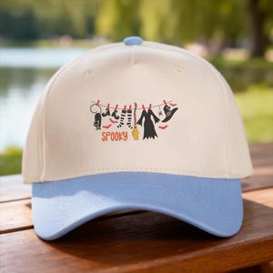 May include: A cream-colored baseball cap with a light blue brim. The front features a graphic of a clothesline with a black cat, striped socks, a broom, a dress, a spider, and a witch hat, with the orange text SPOOKY printed underneath.