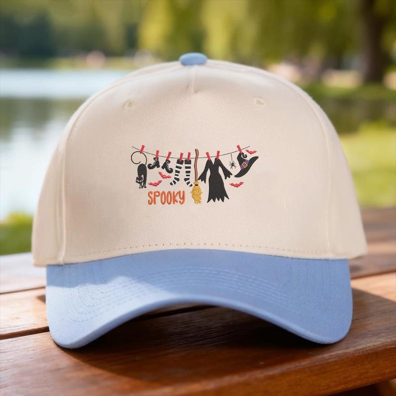 May include: A cream-colored baseball cap with a light blue brim. The front features a graphic of a clothesline with a black cat, striped socks, a broom, a dress, a spider, and a witch hat, with the orange text SPOOKY printed underneath.