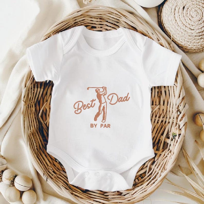 May include: White baby onesie with a brown graphic of a golfer and the words "Best Dad BY PAR". The onesie is displayed in a woven basket, suggesting a gift or baby item.