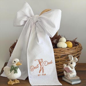 May include: A white golf towel with the text "Best Dad By Par" and a golfer graphic, hanging from a woven basket. The basket contains speckled eggs. Decorative duck and bunny figurines with glasses are on either side.