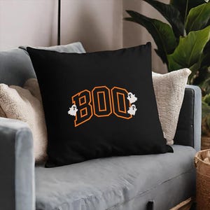 May include: Black square throw pillow with the word "BOO" in orange block letters and three white ghost figures. The pillow is on a gray couch with white and beige pillows.
